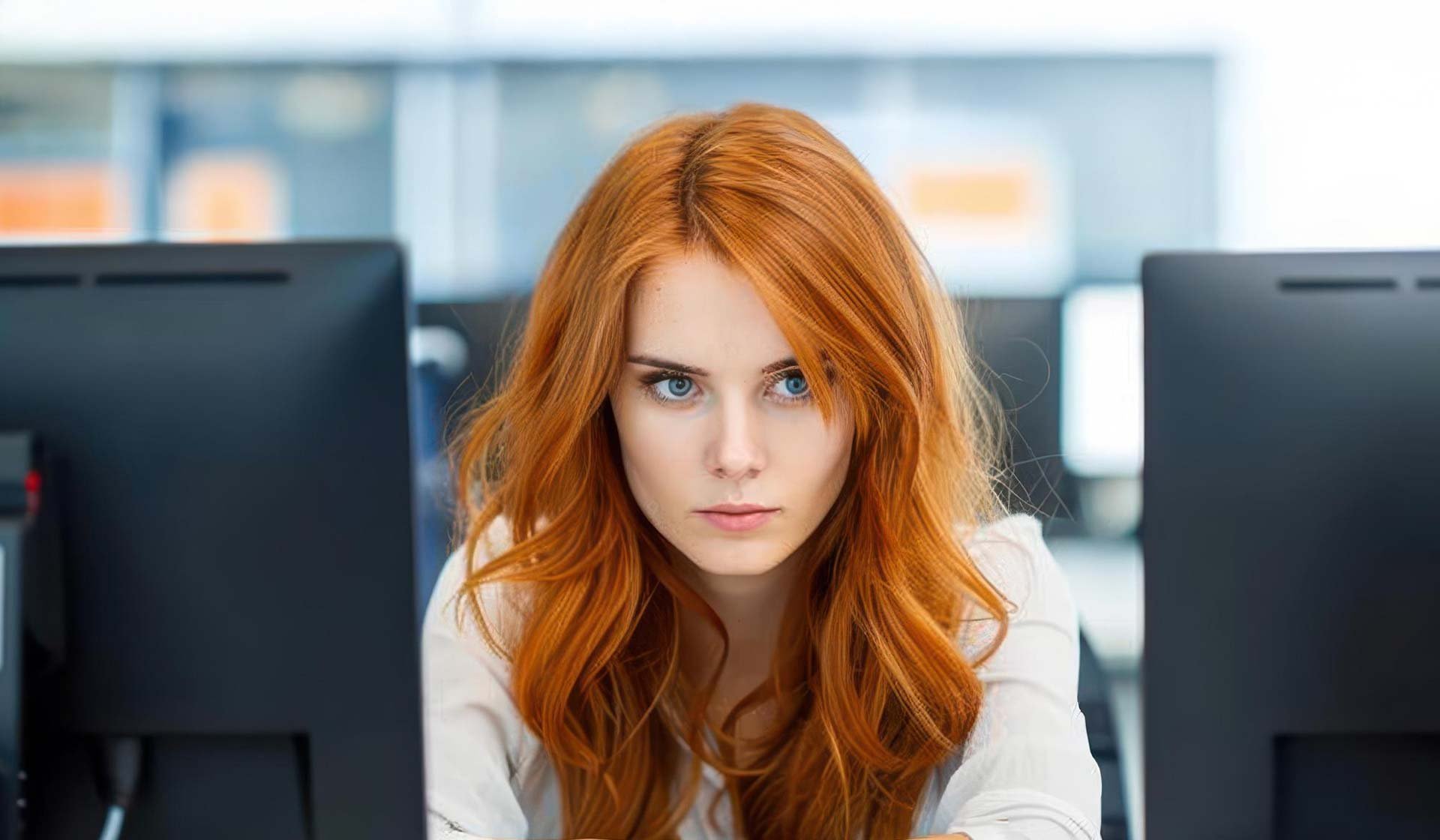 Junge Frau mit langen roten Haaren konzentriert vor zwei Bildschirmen im Büro – Symbolbild für digitale Baulohnabrechnung in der Cloud oder On-Premise