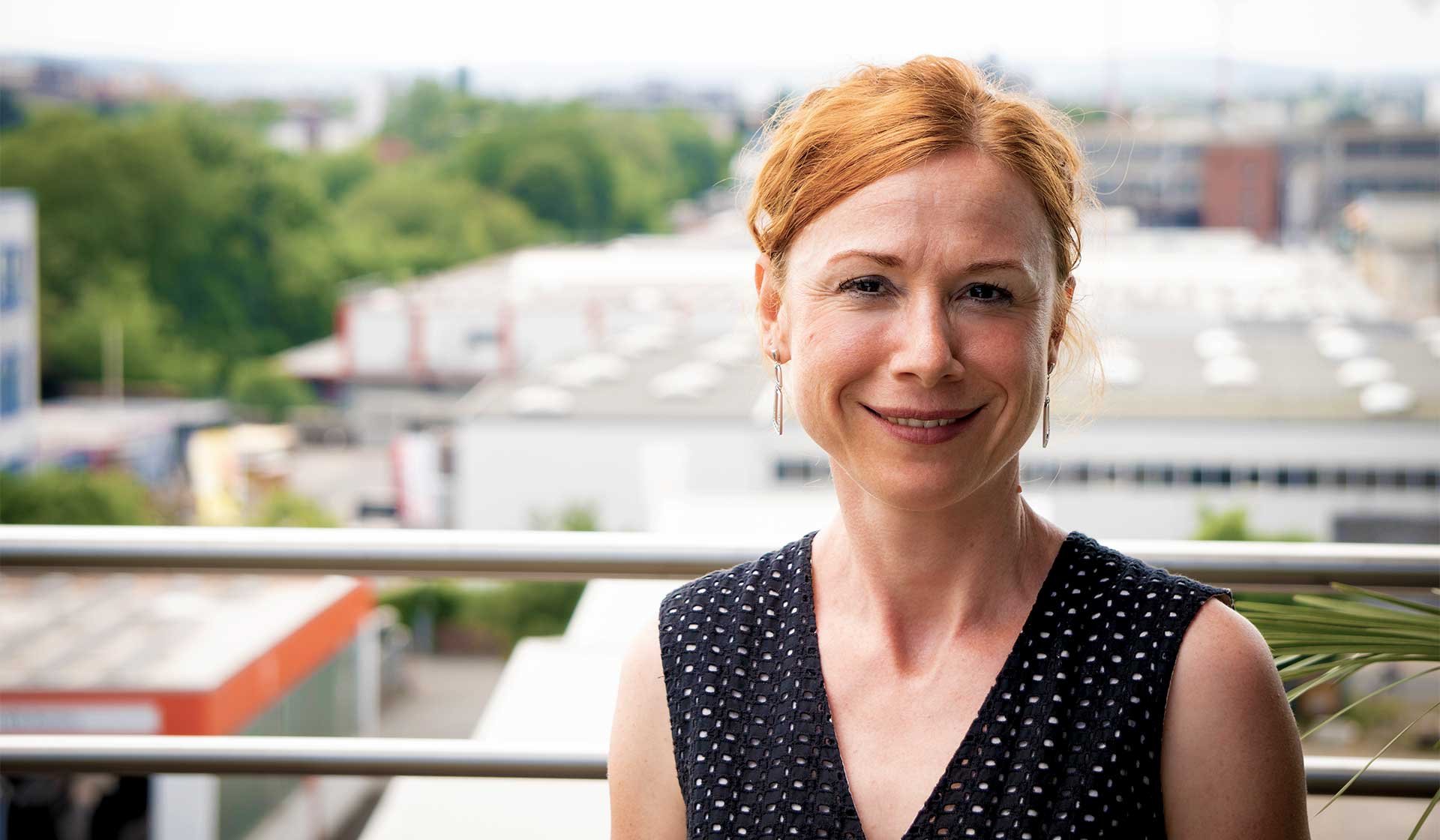 Porträt einer freundlich blickenden Frau auf dem Balkon eines Bürogebäudes mit Stadt- und Industriehintergrund – Symbolbild für ein Experteninterview zur Baulohnabrechnung