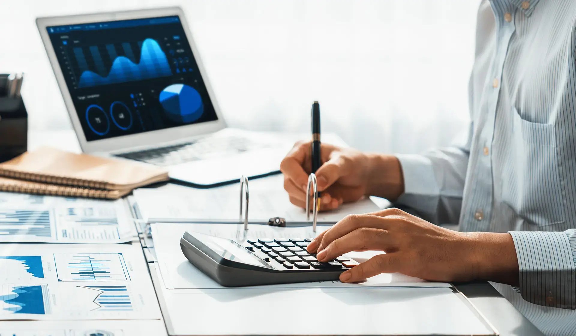 Person rechnet mit Taschenrechner und analysiert Finanzdaten – Diagramme und Tabellen auf dem Schreibtisch sowie auf dem Laptop – Symbolbild für Beitragsbemessungsgrenzen und Lohnabrechnung
