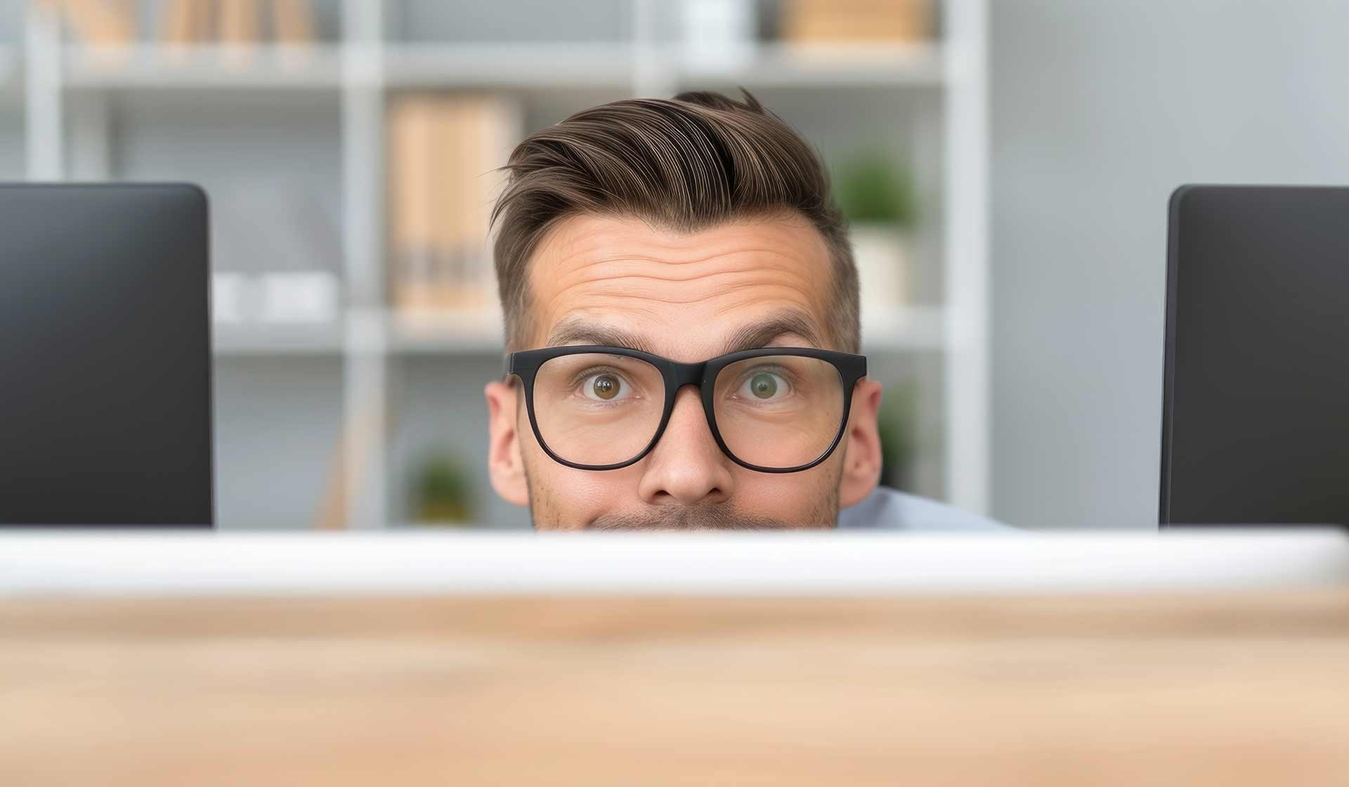 Überraschter Büroangestellter mit Brille blickt hinter einem Bildschirm hervor – symbolisch für unerwartete Erkenntnisse oder aufgedeckte Mythen
