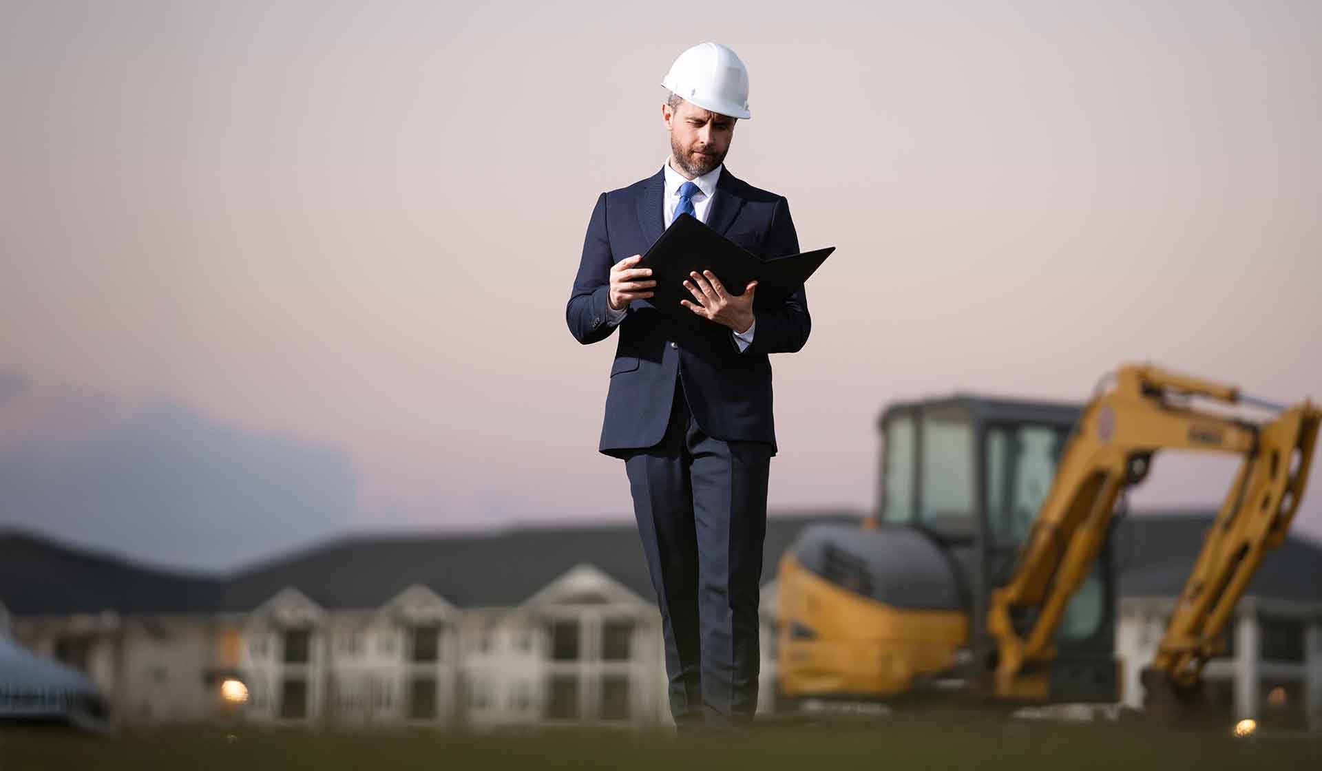 Bauleiter im Anzug mit Schutzhelm prüft Unterlagen auf der Baustelle bei Sonnenuntergang – Symbolbild für betriebswirtschaftliche Umlagen im Bauwesen